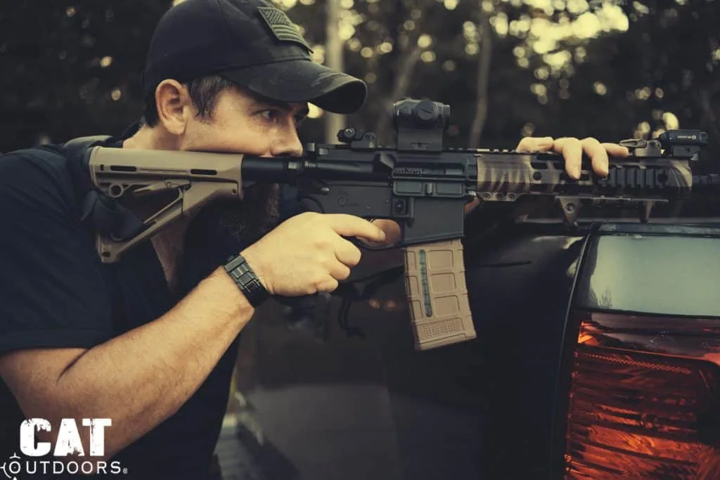 Bearded shooter in a black cap aims a camo AR-15 with a tan stock beside a pickup truck