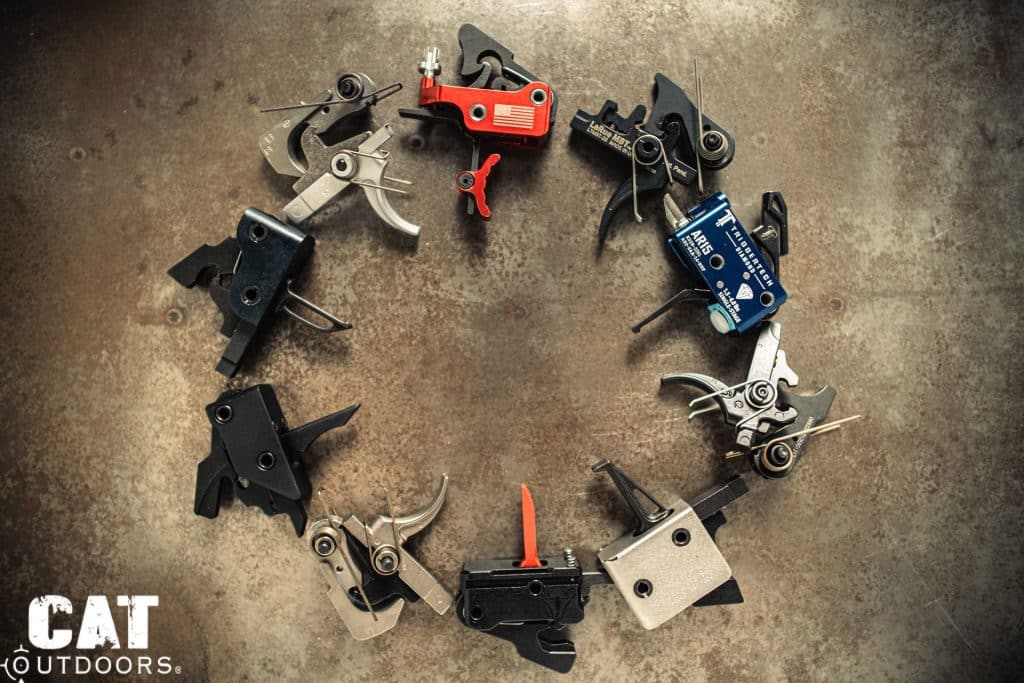Ten AR-15 drop-in triggers arranged in a circle on a weathered steel backdrop, including TriggerTech, LaRue MBT, and competition models