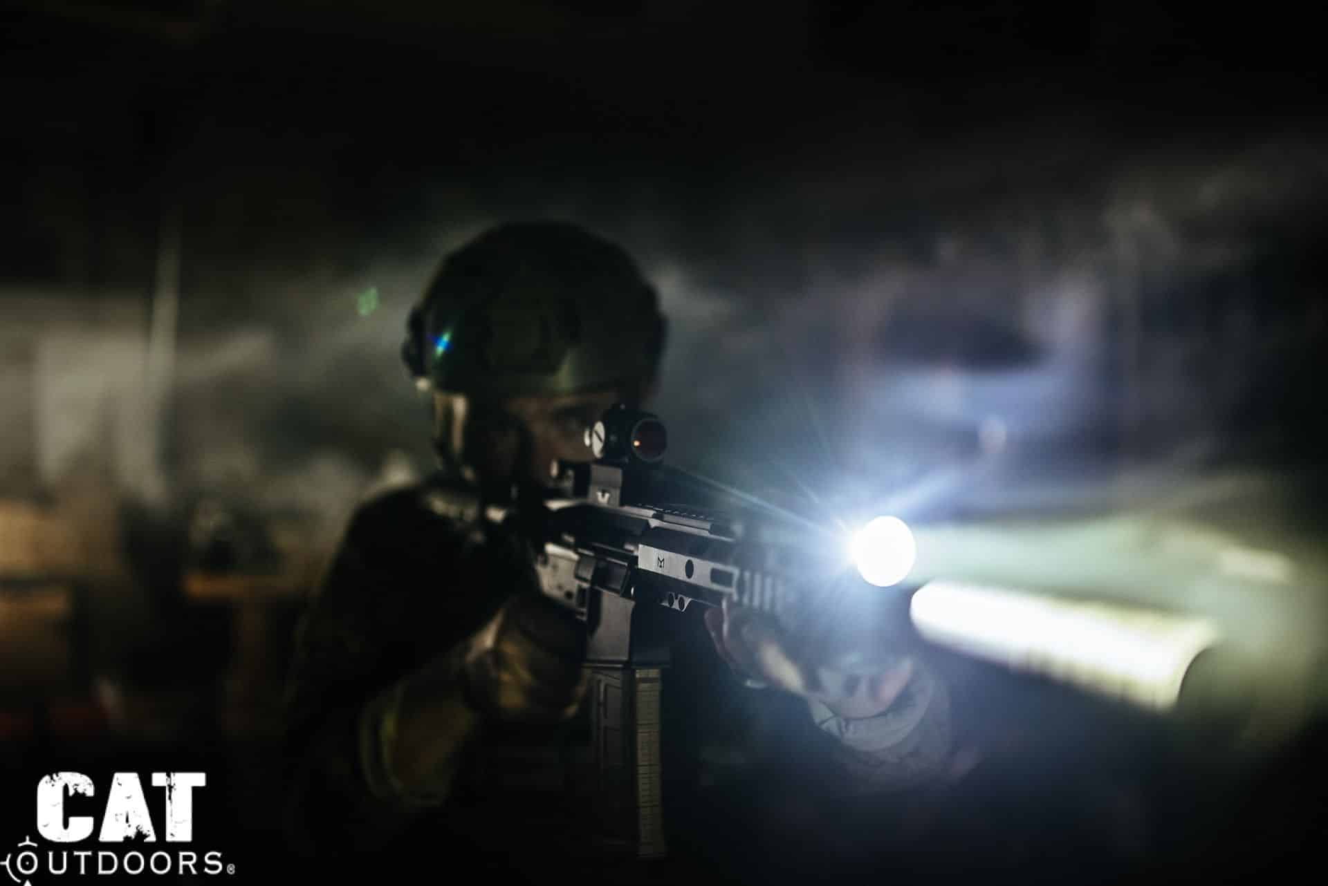 Shooter in helmet and gear aiming an AR-15 with a mounted weapon light illuminating a dark room