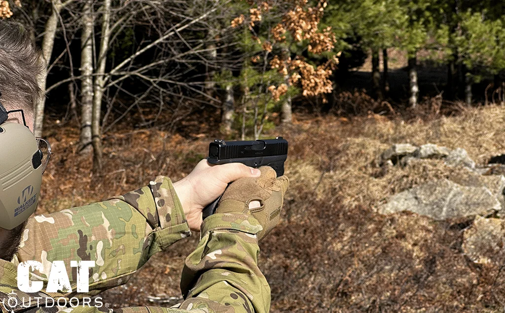 Shooter in multicam and electronic earmuffs aiming the 43X two-handed at a wooded outdoor range