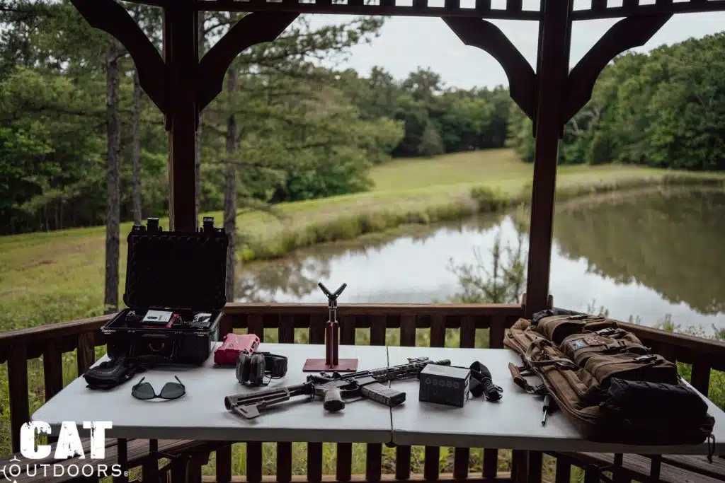AR-15, optic box, ear muffs and an open hard case on a pavilion table overlooking a pond