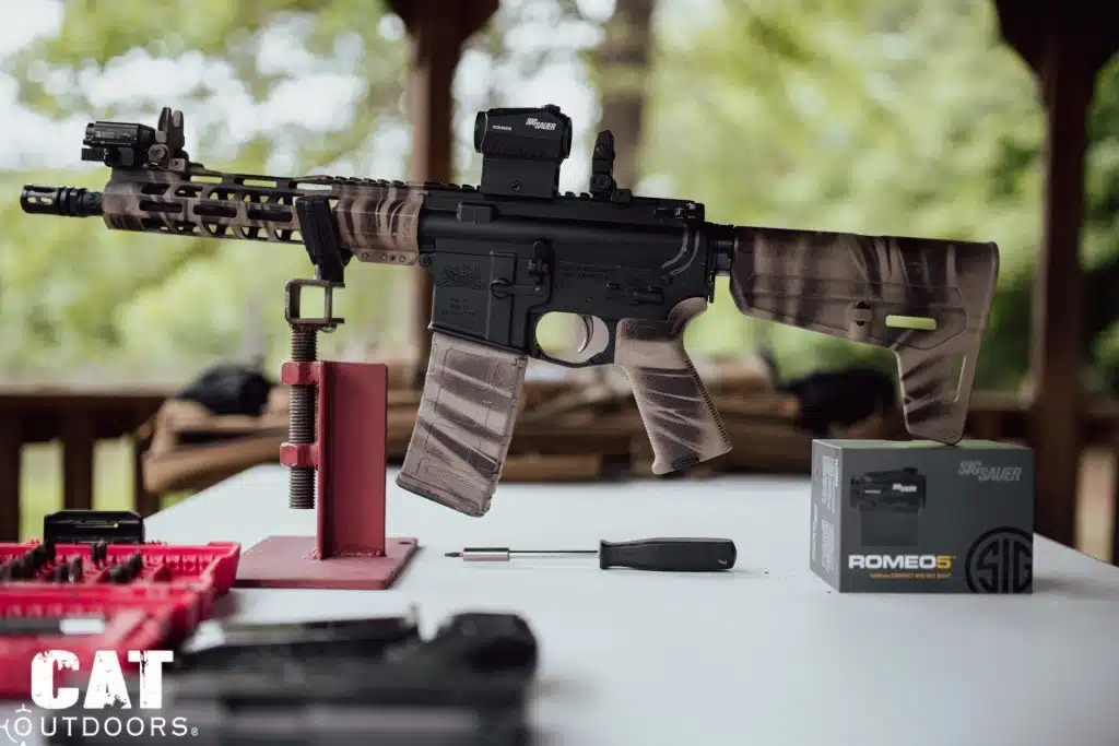 Camo-wrapped AR-15 on a red bench stand, Romeo 5 mounted and its retail box beside the rifle