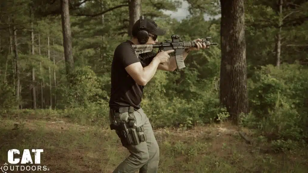 Standing shooter with a drop-leg holster, AR-15 shouldered and eye behind the Romeo 5, in a pine clearing