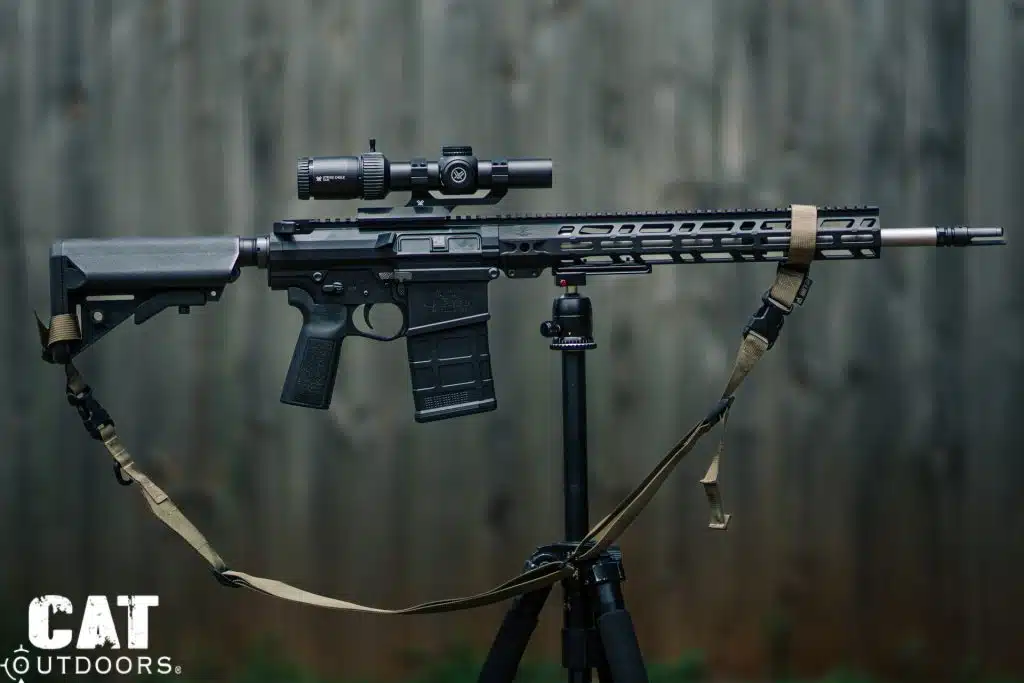 Scoped AR-10 with a tan sling balanced on a tripod in front of a weathered wooden fence