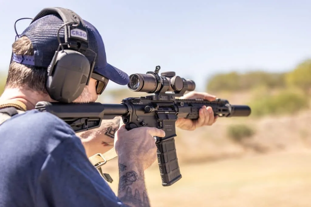 Shooter in a cap and ear protection aims a suppressed AR-15 with an LPVO on a sunny range