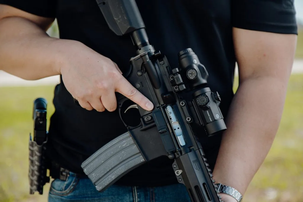 AR-15 held at the chest with a red dot and 3x magnifier, finger indexed along the receiver
