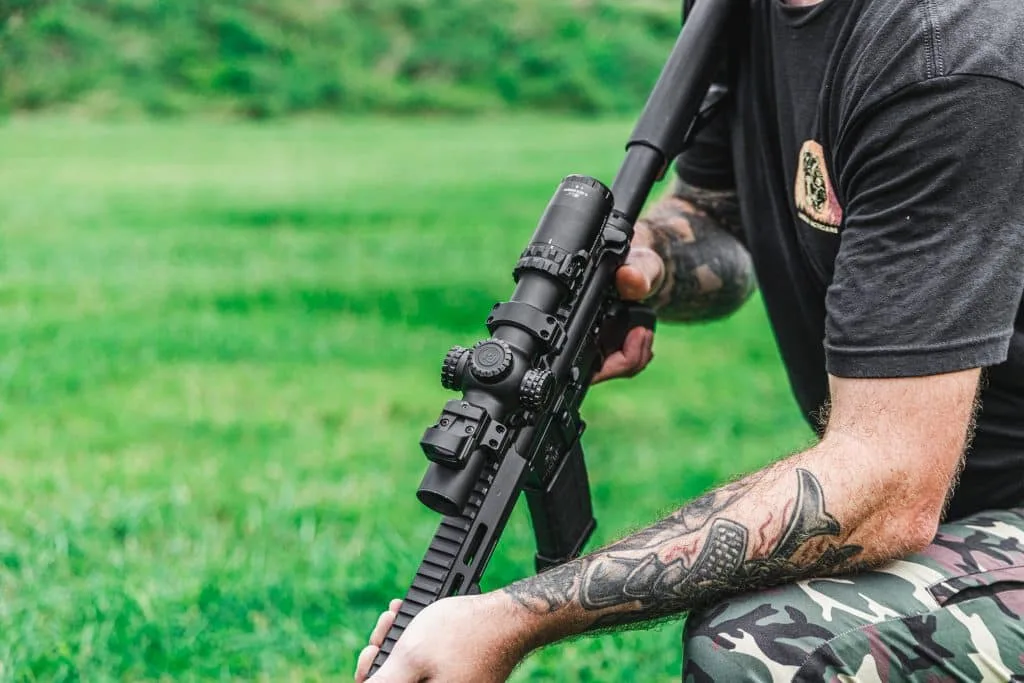 Kneeling shooter holds an AR-15 topped with an LPVO and a mini red dot on the scope mount