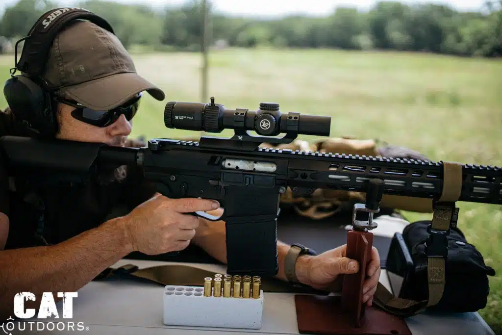 Shooter in ear protection aims a scoped AR-10 from a wooden rest beside a block of brass cartridges