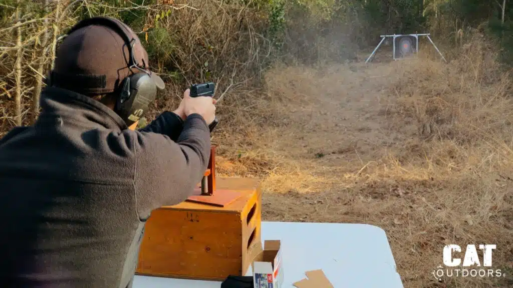 Brad firing the Springfield Hellcat from a seated bench rest position at an outdoor range with a target visible downrange
