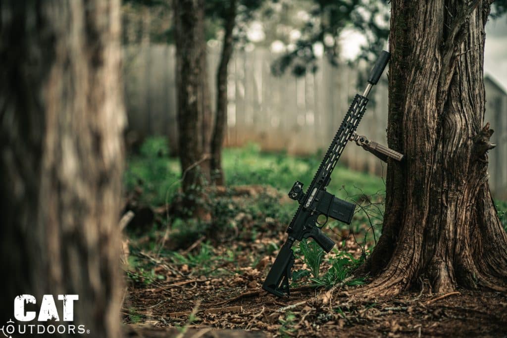Suppressed AR-15 with CVLIFE bipod folded leaning against a tree in wooded setting