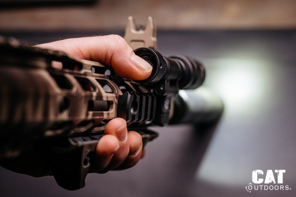 Shooter's hand reaching forward to activate the tailcap from a normal AR-15 firing grip, light beam visible on wall